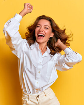 A young woman in a white shirt is exulting and laughing with her right fist raised in the air. She has her left hand on her hair and her head turned to the side.