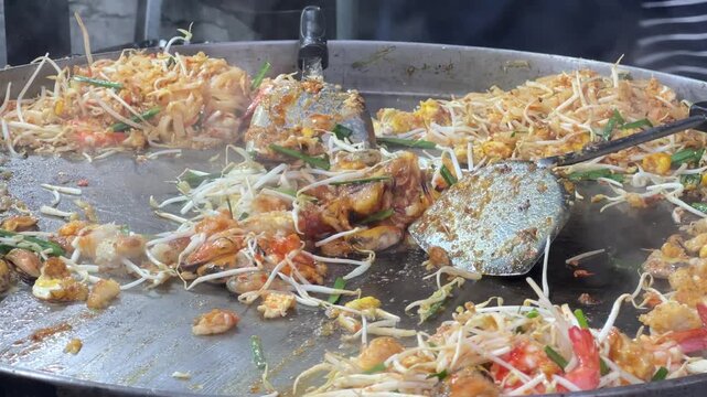 Asian street food vendor using spatulas to stir fry noodles and seafood on big hot pan.