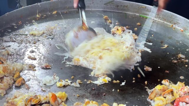Asian street food vendor using spatulas to stir fry noodles and seafood on big hot pan.