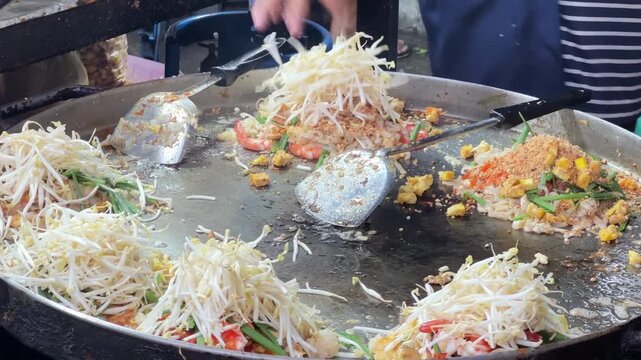 Asian street food vendor using spatulas to stir fry noodles and seafood on big hot pan.