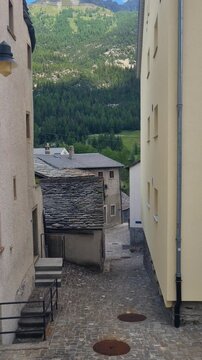Vertical view down a cobblestone street in Simplon Dorf, Switzerland, lined with traditional stone houses and framed by the Alpine landscape, capturing the timeless charm of a historic Swiss village