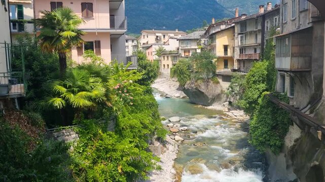 Picturesque view of Chiavenna, Lombardy, Italy, with colorful riverside buildings along the Mera River, Mediterranean vegetation, and the rugged Rhaetian Alps, showcasing this historic alpine village