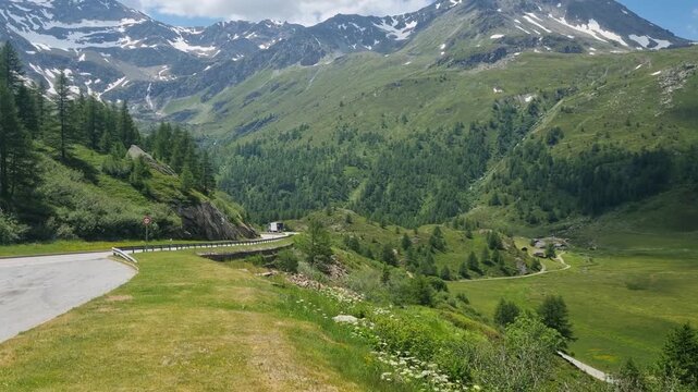 Empty mountain road crossing Simplon Pass in Valais, Switzerland, with partly snow-covered Alpine peaks rising above lush green meadows, capturing a scenic Swiss road trip on a bright summer day