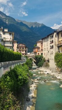 Scenic vertical view of Chiavenna, Lombardy, Italy, the Mera River, lush Mediterranean vegetation, and the dramatic Rhaetian Alps, highlighting the charm of this historic Alpine town