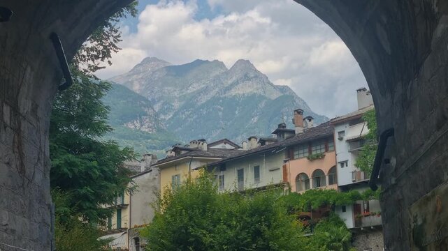 Framed by a stone bridge arch, Chiavenna in Lombardy, Italy, overlooks the Mera River, lush Mediterranean vegetation and the dramatic Rhaetian Alps, blending alpine scenery with historic charm