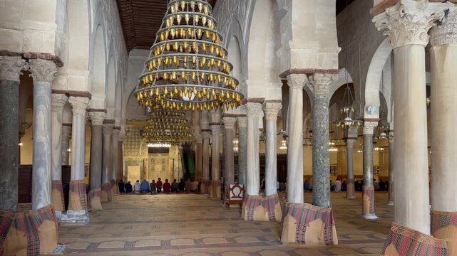 Worshippers sit in rows inside the Great Mosque of Kairouan, framed by marble columns and large chandeliers in a historic prayer hall. Tunisia.