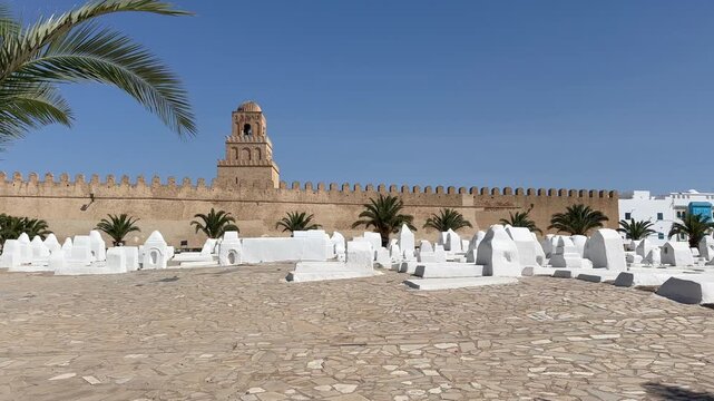 White tombs stand before the fortified wall and tower of the Great Mosque of Kairouan, framed by palm trees on a bright sunny day. Tunisia.