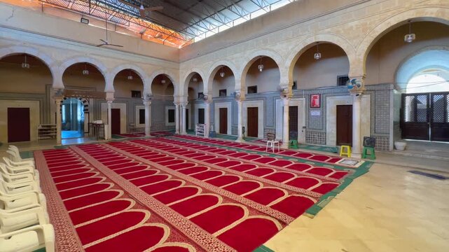 Wide shot of an empty mosque interior in Zawiya of Sidi Sahib, also known as the Zawiya of Abu al-Balawi or Mosque of the Barber, with red prayer carpet, white arches and tiled walls.