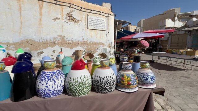 Hand-painted ceramic pots line a table beside a weathered plaster wall in a sunlit Tunisian market scene with stalls in the background.