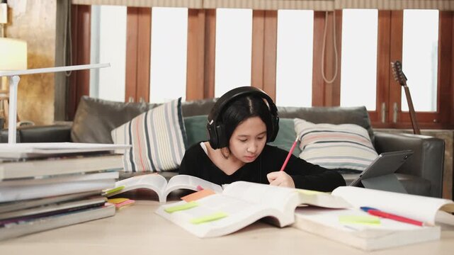 Asian teenage girl in headphones concentrates on learning at home, reading textbooks, note writing, self-tutorial exams, preparation for back to school, Homeschooling, university education lifestyle.