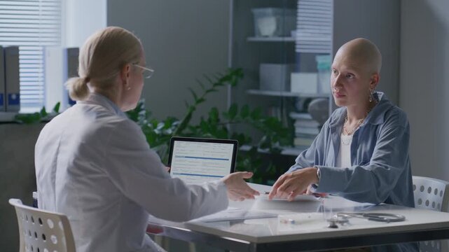 Smiling bald female cancer patient signing medical agreement and shaking hands with supportive doctor during consultation at hospital
