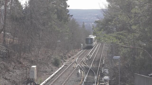 Elevated shot of an Oslo Metro T-bane train traveling along outdoor tracks framed by pine trees on the hillside leading to Frognerseteren station, with distant valley views in Oslo, Norway.