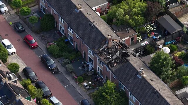 High angle aerial footage revealing the devastating aftermath of a house fire in a residential neighborhood, showing the collapsed and charred roof structure of a terraced building