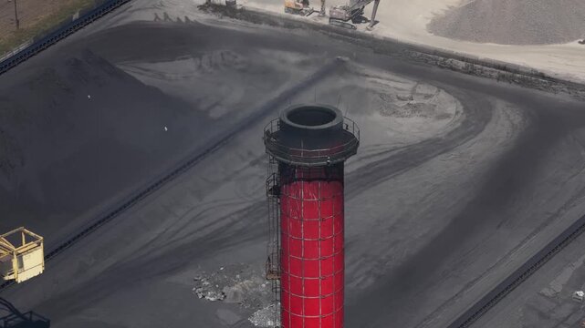 Aerial view of a large coal storage yard featuring a prominent red and white smokestack, with a massive yellow stacker reclaimer machine moving along tracks to manage the fuel pile