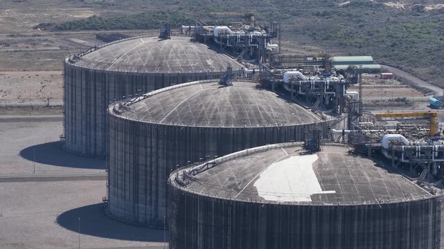 Aerial view of three large industrial lng storage tanks releasing excess pressure through a safety valve, with vapor cloud venting into the atmosphere at a modern energy facility