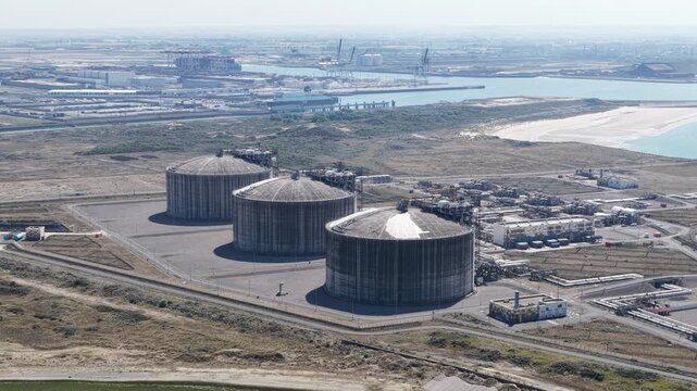 Expansive aerial footage showcasing large liquefied natural gas storage tanks at an industrial plant, with a busy commercial seaport and vast coastline visible in the hazy background