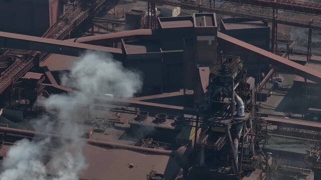 Towering structures of a large metallurgical plant emitting puffs of white smoke, showcasing the immense scale and complexity of heavy industry and manufacturing from a high aerial perspective