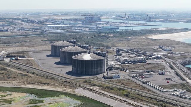 Expansive aerial footage showcasing a liquefied natural gas storage facility with large cylindrical tanks, pipelines, and a processing plant located next to a busy industrial seaport on the coast