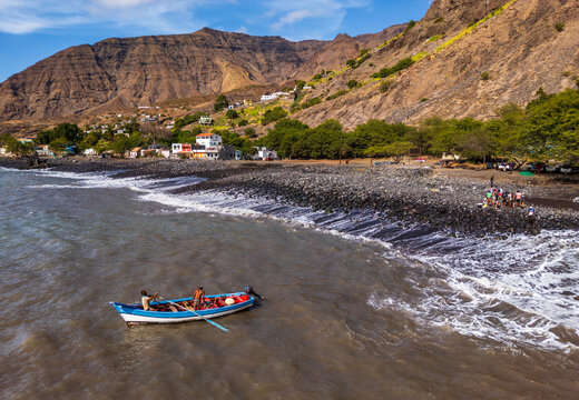 Tarrafal, Cabo Verde - 18 February 2025: Aerial view of a fishing boat with two men on the dark volcanic beach of Tarrafal, with coastal mountains and buildings under a blue sky.