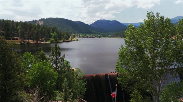 Evergreen Lake dam structure American flag nature landscape downtown Evergreen Colorado aerial drone morning sunny clouds blue sky spring summertime small Rocky Mountain town forward pan up motion