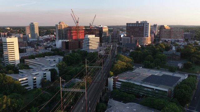 Construction cranes rise above downtown New Brunswick New Jersey at golden sunset, railroad tracks and power lines cut through dense city buildings and green trees below. Aerial drone wide shot