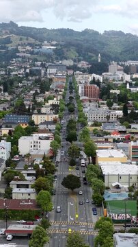Tree-lined boulevard leads toward UC Berkeley Sather Tower amid green hillside in Berkeley California, cars travel busy multi-lane street through dense downtown buildings. Aerial drone vertical shot