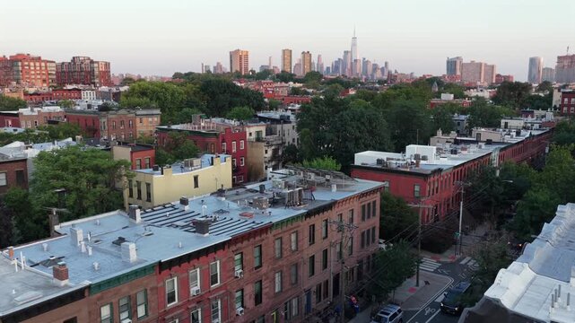 Rows of brick townhouses with flat white rooftops stretch through residential Hoboken, New Jersey, dense tree canopy and Manhattan skyline rise beyond in evening light. Aerial drone passing wide shot