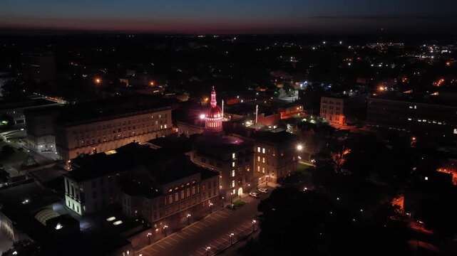 Illuminated gold dome of New Jersey State House rises above downtown Trenton buildings at night, streetlights and city lights spread across surrounding rooftops below. Aerial drone backwards wide shot