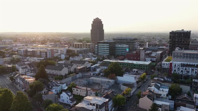 PPL Tower rises above downtown Allentown, Pennsylvania, glass office buildings and brick rowhouses spread through dense city center as golden sunset haze slowly fades. Aerial drone wide shot