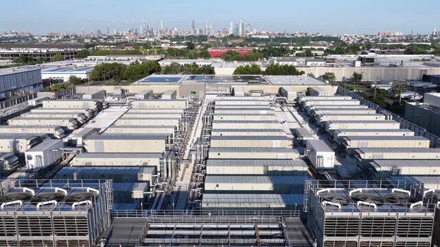 Rows of industrial cooling units and ductwork cover large data center rooftop in Secaucus, New Jersey, distant Manhattan skyline rises beyond flat warehouse buildings. Aerial drone flyover shot