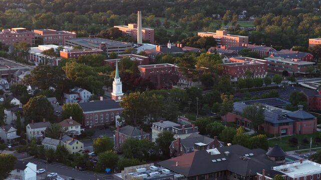 White church steeple with clock rises among red brick institutional buildings in New Brunswick, New Jersey, dense green tree canopy glows warmly at golden morning sunrise. Aerial drone wide shot