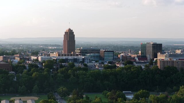 PPL Tower rises above downtown Allentown, Pennsylvania, glass and brick office buildings spread beyond dense green tree canopy under hazy golden morning sunrise light. Aerial drone view.