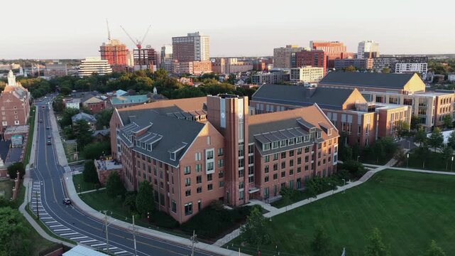 Car drives along curving road past brick dormitory building in New Brunswick, New Jersey, downtown skyline and construction cranes rise beyond green lawns at sunset light. Aerial drone view.