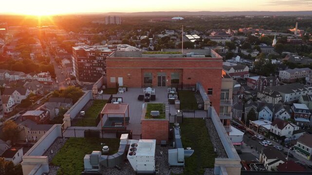 Luxury rooftop terrace with HVAC units and green plantings tops brick building in downtown New Brunswick, New Jersey, dense residential rooftops glow at golden sunset. Aerial drone flyover shot