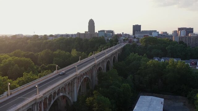 Arched concrete Albertus Meyers Bridge carries cars over dense green treetops toward downtown Allentown, Pennsylvania, PPL Tower rises above city skyline at golden sunset. Aerial drone descend shot