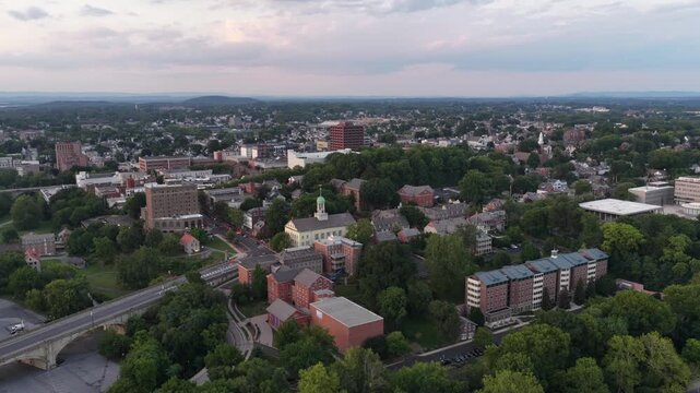 Historic homes with steep gabled roofs peek through summer tree canopy, downtown Bethlehem skyline rises beyond wooded hills in residential Pennsylvania neighborhood. Aerial drone wide shot