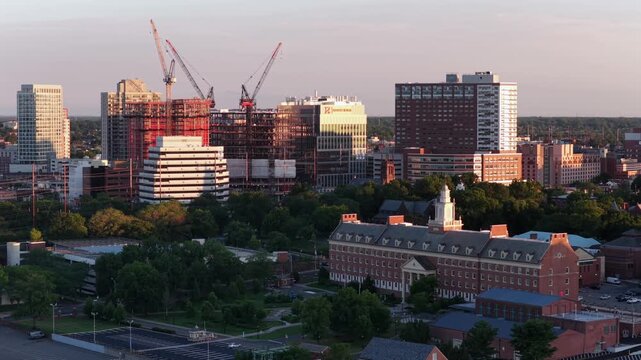 Construction cranes rise above downtown New Brunswick, New Jersey, red brick dormitory building and modern glass towers glow warmly under golden evening sunset light. Aerial drone wide shot