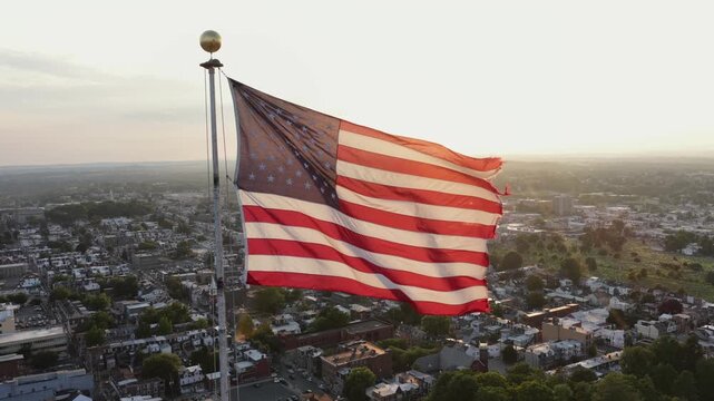 American flag waves atop tall flagpole above dense rowhome rooftops in small American town, warm golden sunset light spreads across quiet residential streets and trees. Aerial drone view.