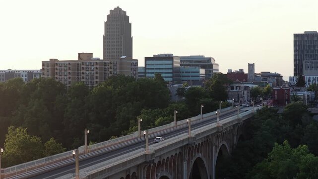 Cars cross historic Albertus Meyers Bridge above dense green treetops in Allentown, Pennsylvania, PPL Tower rises above hazy downtown city skyline at warm golden sunset. Aerial drone rising shot