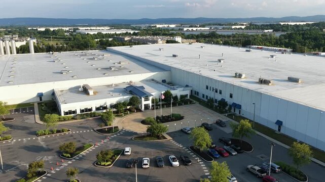 White industrial warehouse spans large distribution park with flat rooftop HVAC units, landscaped entrance and parking lot surround facility near Allentown, Pennsylvania. Aerial drone wide shot orbit