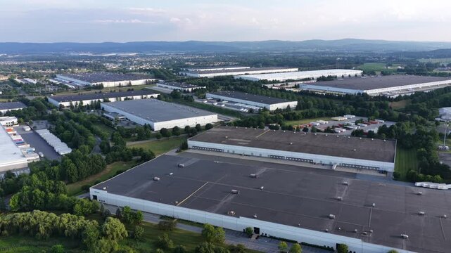 Rows of flat roofed warehouses fill industrial park bordered by trees in Allentown, Pennsylvania, distant mountains rise beyond scattered parking lots and access roads. Aerial drone rising wide shot