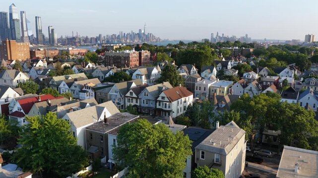 Peaked roof homes and tree canopy fill hillside neighborhood in Union City, New Jersey, Manhattan skyline rises across river beyond waterfront towers in afternoon haze. Aerial drone flyover shot
