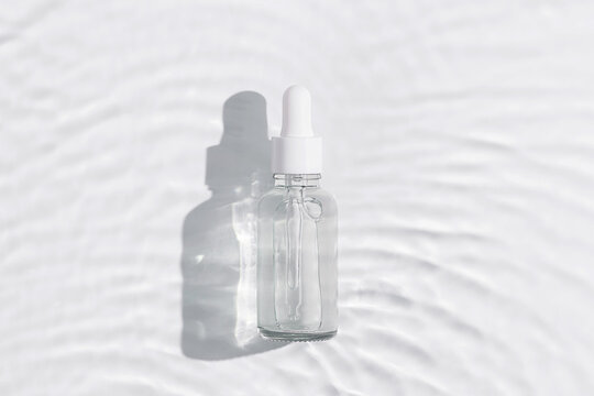 Natural cosmetic facial skin care product in glass bottle with white lid and pipette with colorless liquid essence, serum on waves wanter background, dark shadow. Aesthetic mockup cosmetics, top view.