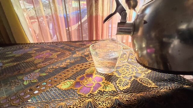 GRESIK, INDONESIA - JULY 30, 2026: Pouring hot water from a kettle into a glass cup on a table with a batik tablecloth.