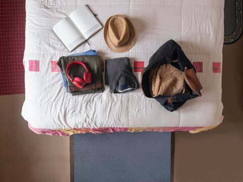 Overhead shot of open travel bag with folded clothes, hat, red headphones, and open notebook on bed