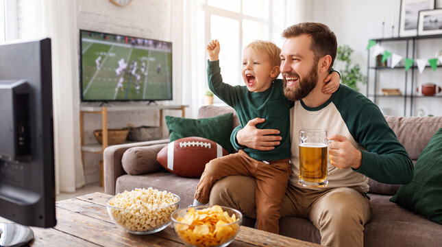 Toddler sits on fathers lap while watching football gameday on television at home with popcorn chips beer for party fun