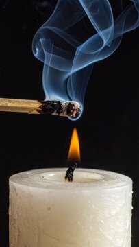 Close up of a burning match lighting a white candle flame with ethereal smoke trails against a dark background