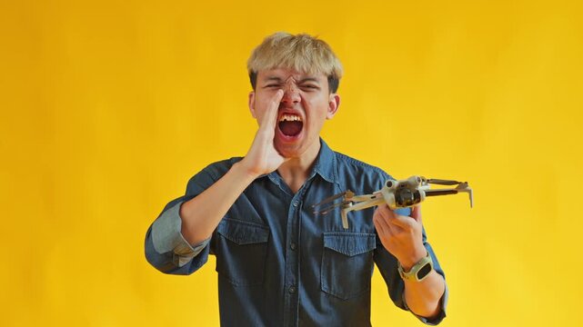 Young man shouting loudly with hand by mouth while holding compact drone on yellow background