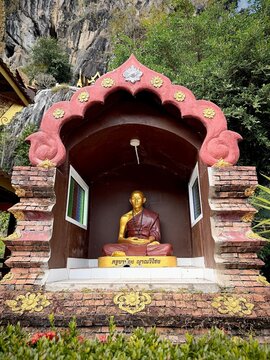A golden monk statue inside an ornate red shrine set against a limestone cliff face at Wat Tham Chetawan in Nan province, Thailand.