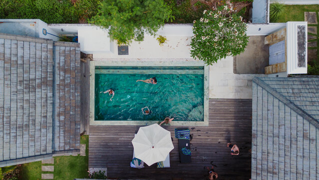 Balikpapan, East Kalimantan, Indonesia - 21 July 2026: Aerial view of a swimming pool at a private villa, with people swimming, a wooden deck, white umbrella, and lush greenery.